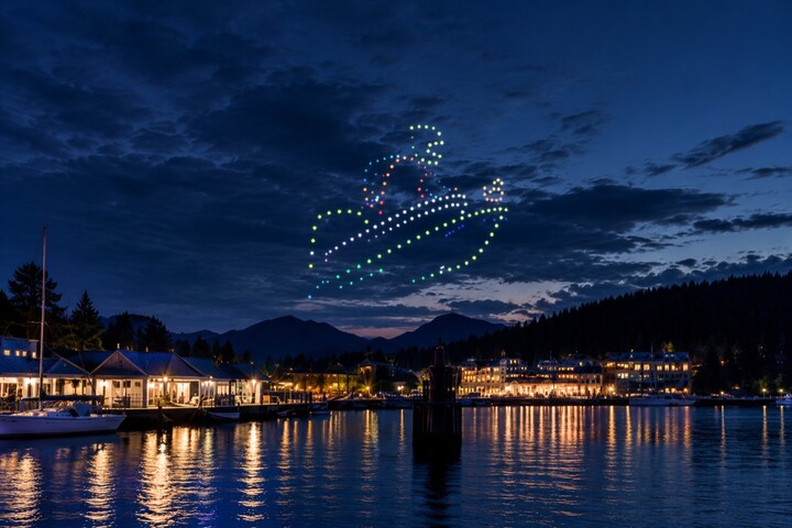Concept: a colourful drone formation above a waterfront venue and marina, with evening lights reflected in the water.